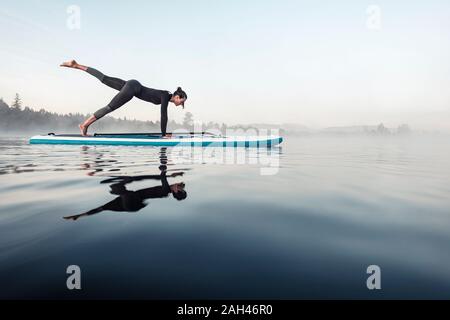 Woman practicing paddle board yoga on lake Kirchsee in the morning, Bad Toelz, Bavaria, Germany Stock Photo