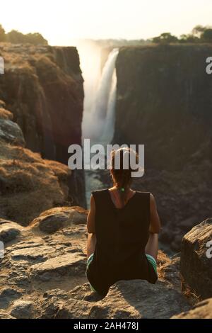 Woman sitting on the top of a rock enjoying the Victoria Falls at sunset, Zimbabwe Stock Photo