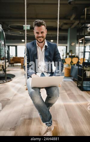 Businessman using laptop in coworking space, sitting on swing Stock Photo