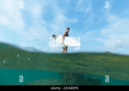 Surfer holding his surf board, standing on stone, Sumbawa island, Indonesia, over-under image Stock Photo