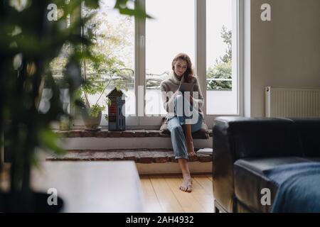 Mature woman sitting on wondow sill, using digital tablet Stock Photo