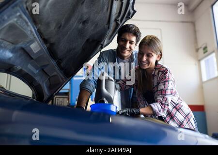Young woman pouring lubricant in funnel while man standing near and watching both working in team in car service Stock Photo