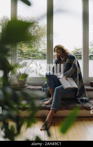 Mature woman sitting on wondow sill, wrapped in blanket, reading book Stock Photo