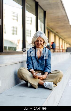 Mature businesswoman sitting cross-legged on steps in the city, using smartphone Stock Photo