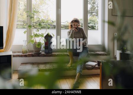 Mature woman sitting on wondow sill, using digital tablet Stock Photo