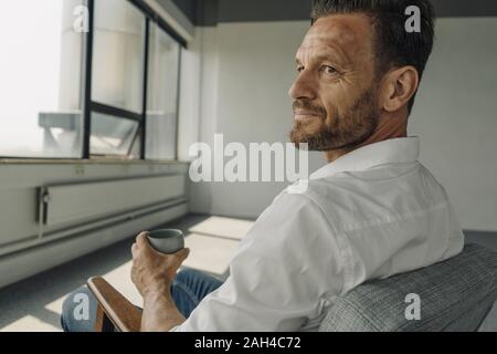 Mature businessman with cup of coffee sitting in armchair in empty office Stock Photo