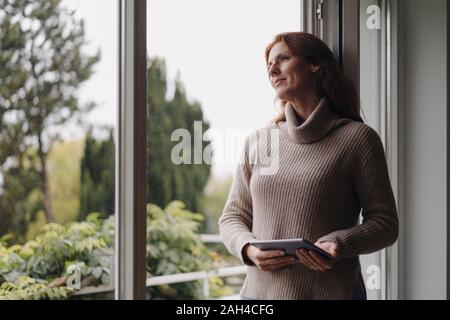 Woman looking out of window, holding digital tablet Stock Photo