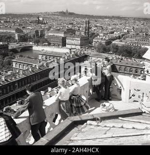 1950s, historical, people standing on top of a building looking over the skyline of the city of Paris, France, with the catholic church of Sacre Coeur de Montmartre standing on the hillside in the far distance. Stock Photo