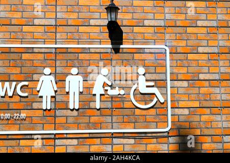 On the orange brick wall is a toilet signpost with white images of  WC, a man, a woman, a baby changing scene of a baby and a person in a wheelchair. Stock Photo