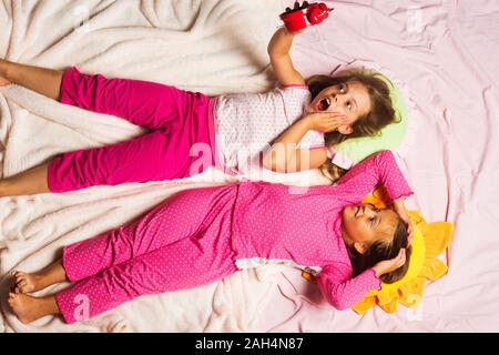 Schoolgirls over slept lying on funny pillows. Children with shocked faces lie on pink background holding alarm clock. Kids in pink pajamas, put hands up. Childhood and deadline concept. Stock Photo