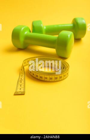 Dumbbells in bright green color isolated on yellow background. Sports equipment and yellow measuring tape in roll. Barbells made of plastic near long soft ruler. Athletics and weight loss concept Stock Photo