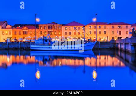 Hobart city harbour named Sullivans cove at sunset with bright illuminated waterfront of historic buildings along pier with fishing boat reflecting in Stock Photo