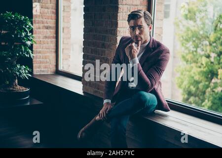 Photo of chic business guy looking clever eyes deeply thinking sitting, window sill hand on chin wear specs stylish blazer turtleneck and jeans in mod Stock Photo