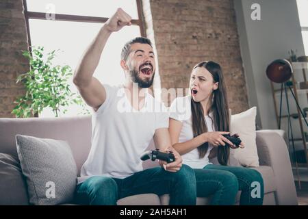 Photo of funny pair, sitting sofa indoors playing video games excited by unexpected winning and losing Stock Photo