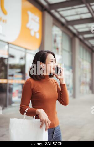 Portrait of happy young Asian woman talking on mobile and carrying paper shopping bags walking outdoor mall Stock Photo