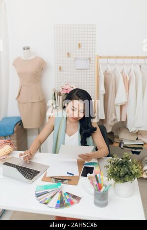 Fashion designer at work. Happy young Asian woman drawing while sitting at her working place in fashion workshop Stock Photo