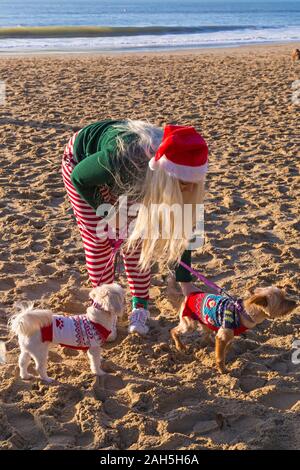 Boscombe, Bournemouth, Dorset, UK. Christmas Day 25th December 2019. Brave volunteers plunge into the sea for a swim or dip on a lovely sunny day, for the annual charity White Christmas Dip, the largest Christmas Dip in the UK, dressed in fancy dress costumes and raising money for Macmillan Caring Locally at Christchurch, a Specialist Palliative Care Unit for patients in the local community. Over a thousand take part in the event which has become a popular tradition for many before their Christmas lunch. Young woman with dogs in their Christmas coats. Credit: Carolyn Jenkins/Alamy Live News Stock Photo