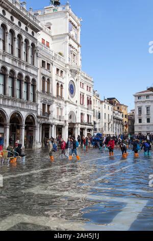 Acqua Alta flooding during extreme high tides Piazza San Marco, Venice, Italy with tourists in colorful boots wading in flood water near Clock Tower Stock Photo