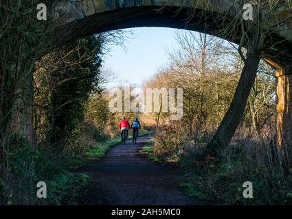 East Lothian, Scotland, United Kingdom, 25th December 2019. Christmas day exercise on the Haddington to Longniddry railway path with a couple riding bicycles seen through the arch of a bridge Stock Photo