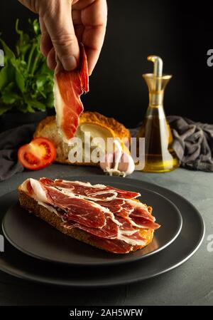 Toasted bread with Iberian ham on black plates and rustic background, a hand holding a slice of ham Stock Photo