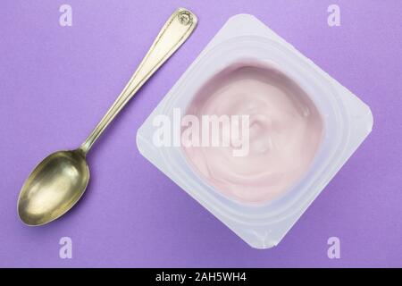 Healthy strawberry fruit flavored yoghurt with natural coloring in plastic cup isolated on purple background with small silver spoon - top view Stock Photo