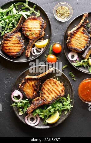 Grilled pork steaks with vegetable leaves on plate over black stone background. Top view, flat lay Stock Photo