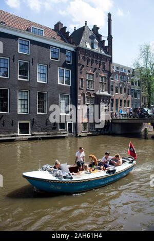View of young people riding a small, blue, open boat in canal doing a tour at Armbrug bridge area in Amsterdam. Historical, traditional buildings are Stock Photo
