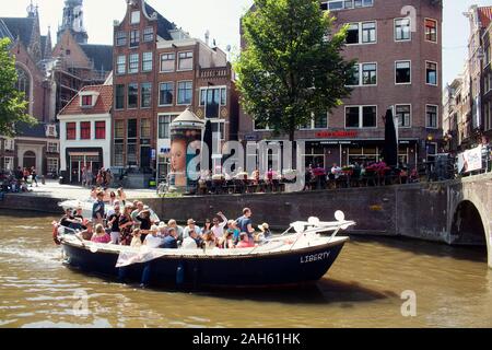 View of people riding a boat on canal doing a cruise tour in front of 13th-century The Oude Church in Amsterdam. Many people hang out on street. It is Stock Photo