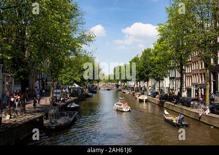 View of people riding small boats on canal doing a cruise tour in Amsterdam. Cloudy blue sky and trees are also in the view. It is a sunny summer day. Stock Photo
