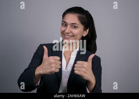 Portrait of a thirty beautiful young business lady in a business suit, showing thumbs up, class, yes, smiling. On a gray background, studio photograph Stock Photo