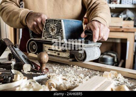 Electric belt sander, sanding machine in male hand. Processing of workpiece on light brown wooden table. Side view, close up Stock Photo