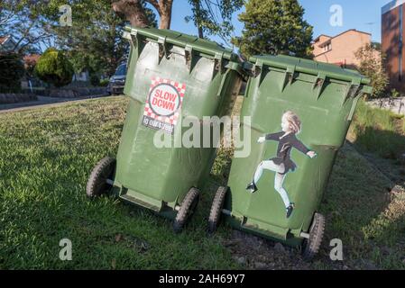 Two large 240L green waste bins with road safety concept stickers or decals attached sit on a roadside footpath facing the morning sun in Sydney, Aust Stock Photo