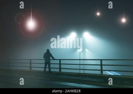 A hooded figure, standing with back to camera on a bridge, looking at UFO alien spaceships coming downfrom the sk., Street lights. On a foggy night. Stock Photo