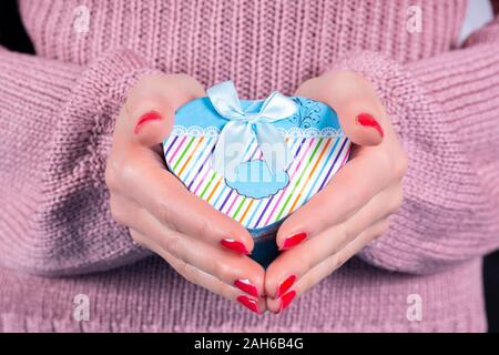 Female hands holding a blue gift box in heart shape and pink woolen sweater in background. Female hands with red nails polish on fingers Stock Photo