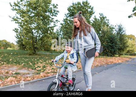 Woman mom teaches son to ride a bike, support care and training, little boy 3-5 years old, summer in city, autumn, emotions of love, care, relaxation Stock Photo
