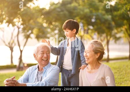 asian grandson, grandfather and grandmother sitting chatting on grass outdoors in park at dusk Stock Photo