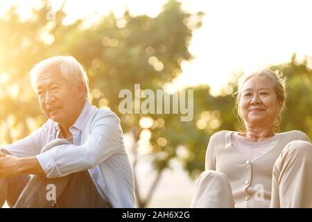 asian senior couple sitting on grass enjoying sunset outdoors in park Stock Photo