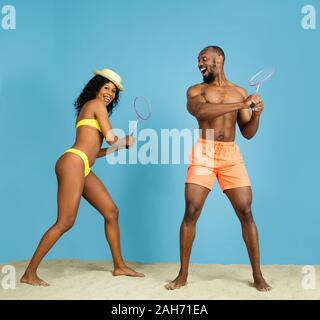 Craziest fun. Happy young african-american couple playing badminton on blue studio background. Concept of human emotions, facial expression, summer holidays or weekend. Chill, summertime, sea, ocean. Stock Photo