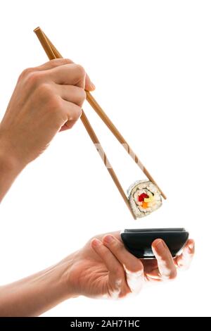 Female hands holding a freshly prepared vegan maki sushi roll with wooden chopsticks and dipping it into a black ceramic bowl with soy sauce - cutout Stock Photo