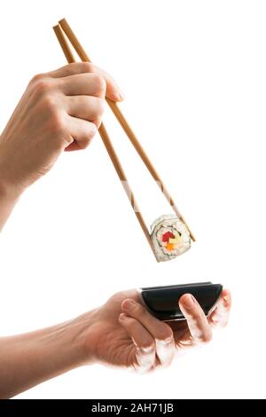 Female hands holding a freshly prepared vegan maki sushi roll with wooden chopsticks and dipping it into a black ceramic bowl with soy sauce, back lit Stock Photo