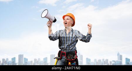 Expressive woman in safety helmet shouting into megaphone. Portrait of young emotional construction worker with loudspeaker on background of modern ci Stock Photo