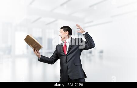 Expressive businessman holding open book. Portrait of adult man in business suit and tie standing in blurred office interior. Excited manager shouting Stock Photo