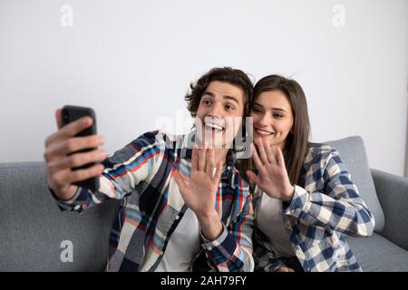 Smiling young couple waving hands while taking selfie on sofa. Stock Photo
