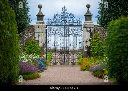 Close up of an ancient iron wrought gate leading into an alleyway lined with colorful bushes of flowers and green hedges Stock Photo