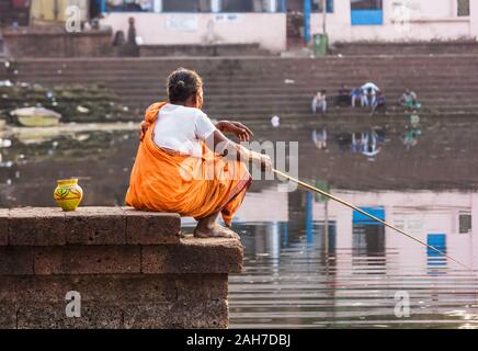 Bhubaneshwar, Orissa, India - February 12 2018: An old woman fishes with a fishing pole while sitting on a stone platform on the ancient Bindu Sagar l Stock Photo
