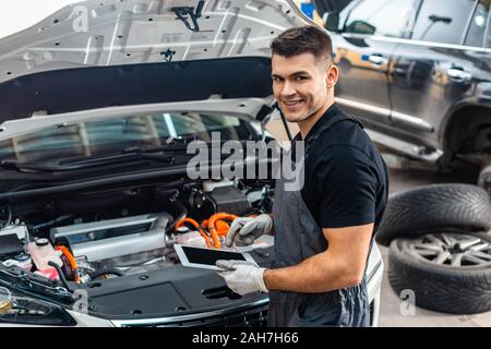 handsome mechanic smiling at camera while using digital tablet near car engine compartment Stock Photo