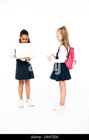 full length view of two multicultural schoolgirls using laptop and digital tablet on white background Stock Photo