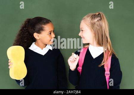 two cheerful multicultural schoolgirls looking to each other while standing near chalkboard Stock Photo
