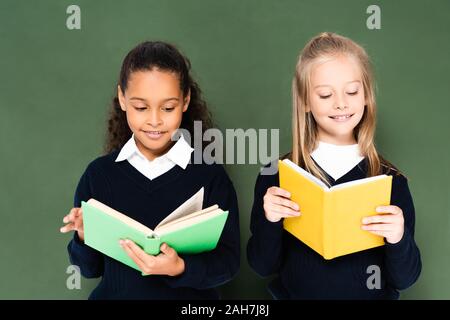 two smiling multicultural schoolgirls reading books while standing near green chalkboard Stock Photo