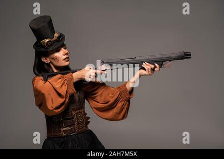 focused steampunk woman in top hat aiming with gun isolated on grey Stock Photo